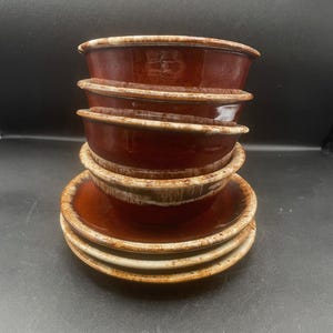 May include: A set of five brown ceramic bowls and saucers. The bowls are a deep reddish-brown with a lighter, speckled rim. The saucers are a similar colour with a matching speckled edge. The set is arranged on a dark background.