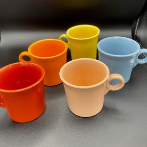 Könnte beinhalten: Fünf Keramikbecher in verschiedenen Farben: Rot-Orange, Orange, Gelb, Hellblau und Pfirsich. Jeder Becher hat einen abgerundeten Henkel und einen leicht ausgestellten Rand. Die Becher stehen auf einer dunklen Oberfläche.