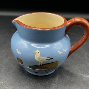 Op de afbeelding: Een blauwe keramische kan met een bruine handgreep. De kan heeft een geschilderd ontwerp van een meeuw op een rots.
