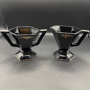 May include: Two black glass creamer and sugar bowls with gold accents and a geometric design. The bowls have a hexagonal base and a wide, rounded top.