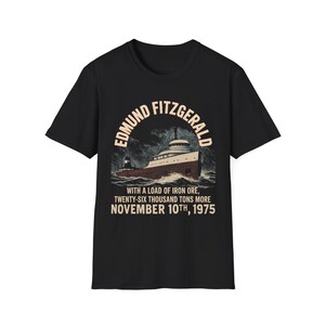 May include: Black t-shirt featuring a vintage illustration of the Edmund Fitzgerald ship. The design includes the ship's name, a depiction of the vessel at sea, and the text "WITH A LOAD OF IRON ORE, TWENTY-SIX THOUSAND TONS MORE NOVEMBER 10TH, 1975."