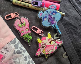 May include: Three acrylic keychains featuring illustrated characters: a pink rabbit with green slime, a blue bear with a zipper mouth, and a pink and white cow with an eye patch. Each keychain includes a pink or purple plastic lobster clasp.