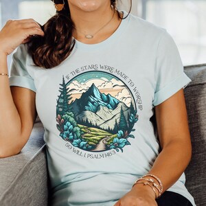 Puede incluir: Una camiseta azul claro con un gráfico de un paisaje de montaña y el texto "Si las estrellas fueron hechas para adorar, yo también lo haré Salmo 148:3".