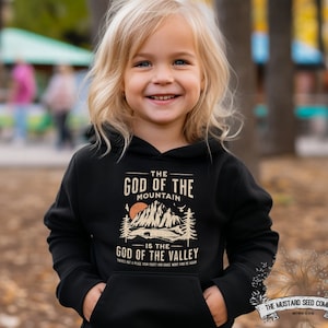 Puede incluir: Una sudadera con capucha negra con un estampado gr&aacute;fico de una cordillera y el texto "The God of the Mountain is the God of the Valley".