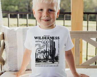 May include: A white t-shirt featuring a black and white desert landscape design with text that reads "Into the Wilderness with God." The shirt has a crew neck and short sleeves. The design includes a desert scene with cacti and the Bible verse Deuteronomy 32:10.