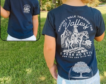 May include: Navy blue short-sleeved t-shirt with a white graphic print of a person riding a horse through a valley. The text "Though I walk through the Valley I fear no evil for you are with me" is printed on the back of the shirt. The text "Psalm 23:4" is also printed on the back of the shirt.