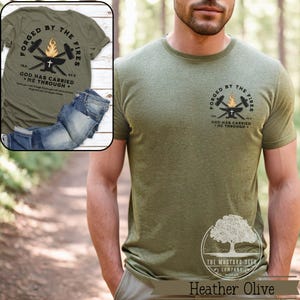 May include: Heather olive green t-shirt with the text "FORGED BY THE FIRES" and "GOD HAS CARRIED ME THROUGH." The shirt features a graphic of a cross, hammer, and flame. The Mustard Seed Company logo is at the bottom.