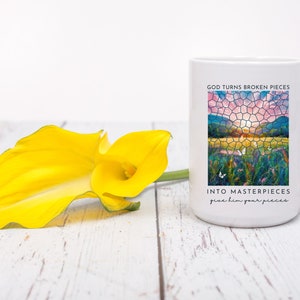 Pu&ograve; includere: Tazza in ceramica bianca con un'immagine colorata in stile vetrata di un tramonto su un campo. Il testo "God turns broken pieces into masterpieces give him your pieces" &egrave; stampato sulla tazza.