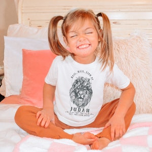 May include: A young child wearing a white t-shirt with a black and white lion graphic and the text "Hail, Hail, Lion of Judah, Let the Lion Roar". The child is sitting on a bed with a white and pink blanket.