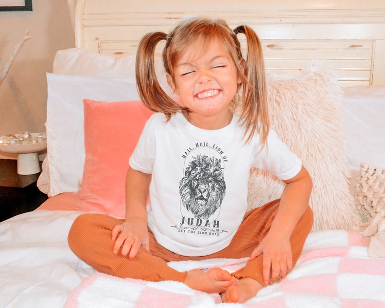 May include: A young child wearing a white t-shirt with a black and white lion graphic and the text "Hail, Hail, Lion of Judah, Let the Lion Roar". The child is sitting on a bed with a white and pink blanket.