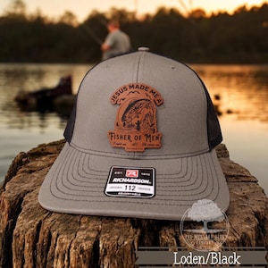 May include: A gray and black trucker hat with a brown leather patch that says "Jesus Made Me A Fisher of Men". The patch features a fish and a person fishing. The hat is adjustable and has a Richardson 112 logo.