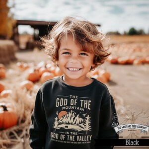 May include: A young boy wearing a black sweatshirt with a graphic design that reads "The God of the Mountain is the God of the Valley." The design features a mountain range, a sunset, and a bear. The boy is smiling and standing in a field of pumpkins.