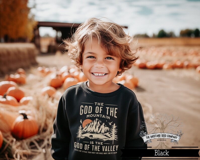 May include: A young boy wearing a black sweatshirt with a graphic design that reads "The God of the Mountain is the God of the Valley." The design features a mountain range, a sunset, and a bear. The boy is smiling and standing in a field of pumpkins.