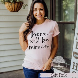 May include: Soft pink t-shirt with the phrase "there will be miracles" in black script. The Mustard Seed Company logo is visible. The shirt has rolled-up sleeves and is paired with blue jeans.