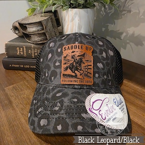 May include: Black leopard print trucker hat with a brown leather patch featuring a cowboy on a horse with the text "Saddle Up" and "Following the Lord". The hat has a black mesh back and a curved brim.