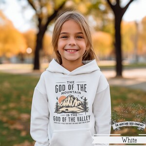 Puede incluir: Sudadera blanca con un dise&ntilde;o gr&aacute;fico que presenta una cordillera y el texto "The God of the Mountain is the God of the Valley". Tambi&eacute;n se incluye el texto "There's not a place your heart and grace won't find the mountain". El logotipo de The Mustard Seed Company est&aacute; en la esquina inferior derecha.