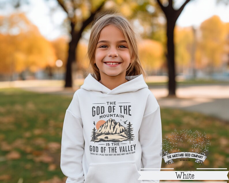Puede incluir: Sudadera blanca con un dise&ntilde;o gr&aacute;fico que presenta una cordillera y el texto "The God of the Mountain is the God of the Valley". Tambi&eacute;n se incluye el texto "There's not a place your heart and grace won't find the mountain". El logotipo de The Mustard Seed Company est&aacute; en la esquina inferior derecha.