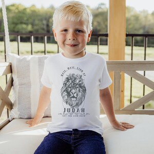 May include: A young boy wearing a white t-shirt with a black and white lion graphic and the text "Hail, Hail, Lion of Judah Let the Lion Roar".