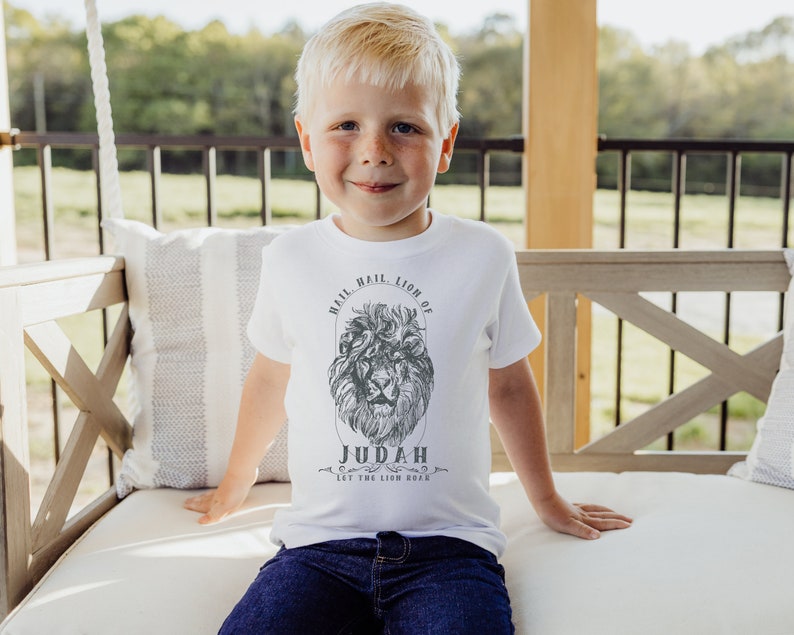May include: A young boy wearing a white t-shirt with a black and white lion graphic and the text "Hail, Hail, Lion of Judah Let the Lion Roar".