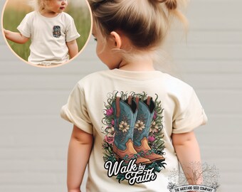 May include: A light beige toddler t-shirt with a graphic of two blue cowboy boots with floral accents. The text "Walk by Faith" is printed below the boots. The t-shirt is made by The Mustang Seed Company.