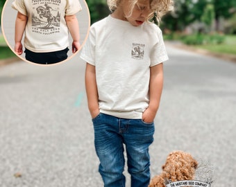 May include: A young child wearing a white t-shirt with a brown graphic of a cowboy riding a horse. The graphic includes the text "Following Jesus Ain't an Easy Road" and "The Only Road Worth Following". The child is wearing blue jeans and standing on a paved road. A small brown dog is sitting in front of the child. The dog is wearing a collar with a tag that says "The Mustard Seed Company". The text "Natural" is printed below the dog.