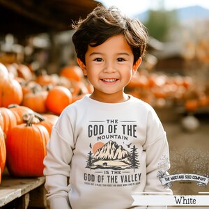 May include: A young boy wearing a white sweatshirt with a graphic design that reads "The God of the Mountain is the God of the Valley." The design features a mountain range, a bear, and a sunset. The boy is standing in front of a pile of pumpkins.