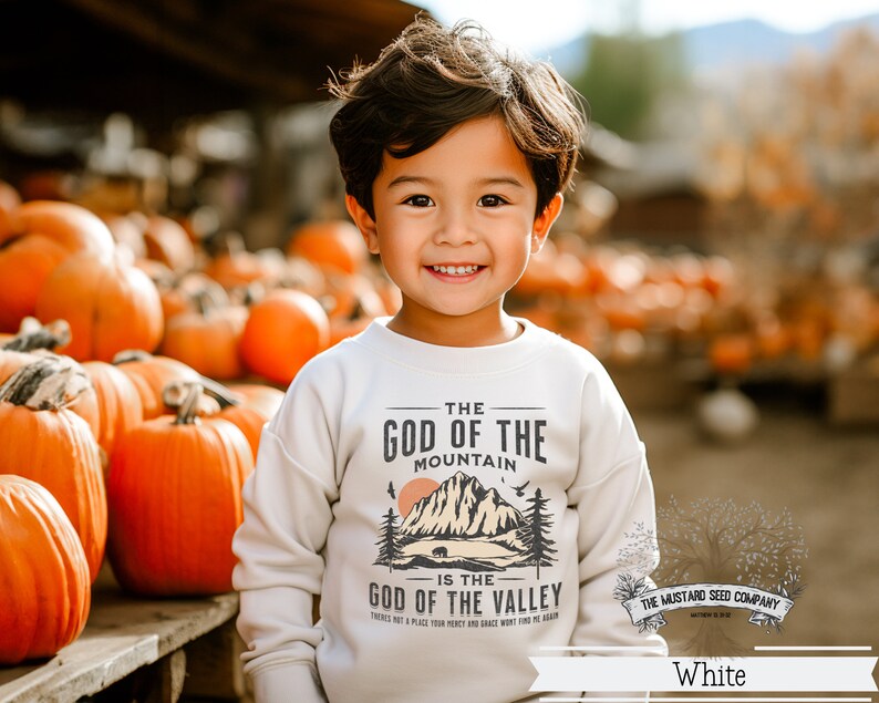 May include: A young boy wearing a white sweatshirt with a graphic design that reads "The God of the Mountain is the God of the Valley." The design features a mountain range, a bear, and a sunset. The boy is standing in front of a pile of pumpkins.