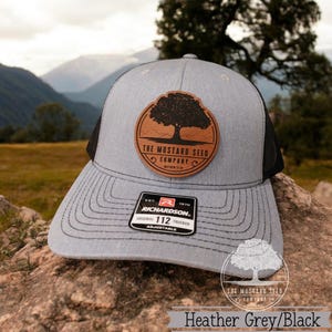 May include: A heather grey and black trucker hat with a brown leather patch featuring a tree and the text "The Mustard Seed Company".