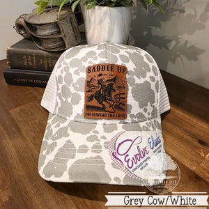May include: A grey and white cow print baseball cap with a white mesh back. The cap features a brown leather patch with a cowboy riding a horse and the text "SADDLE UP" and "FOLLOWING THE LORD".