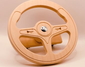 May include: A wooden steering wheel toy with a light brown finish. The wheel has a center hub with a silver metal button and three spokes with a horn design.