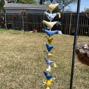 May include: A decorative hanging mobile with fabric bird ornaments in shades of yellow, blue, and white. The birds are suspended from a string, suitable for outdoor display in a garden or yard.