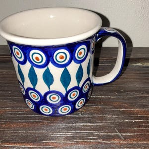Puede incluir: Una taza de cerámica blanca con asa azul y borde ancho. La taza está decorada con un patrón repetitivo de círculos azules con centros naranjas y formas de lágrima turquesa. La taza está sobre una superficie de madera oscura.