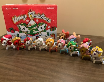 May include: A collection of Tokidoki x Pop Mart Unicorno Christmas figurines, each with unique holiday-themed designs. The figurines are displayed in front of a red box with "Merry Christmas" text and festive illustrations.