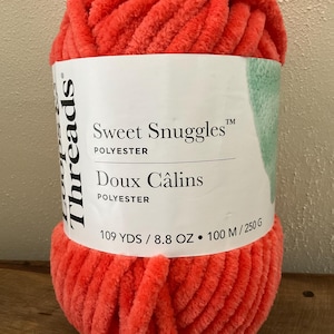 以下が含まれることがあります： 白いラベルが巻かれた、サンゴ色の大きな毛糸玉。「Sweet Snuggles」と「Doux Câlins」と書かれています。糸は100メートル、250グラムです。