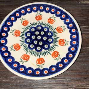 May include: Round ceramic plate with a white base, featuring a blue and orange floral design. The rim has a blue border with orange accents. The center has a cluster of blue dots surrounded by green leaves and orange flower shapes.