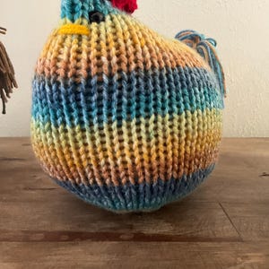 Könnte beinhalten: Eine handgestrickte Hahnfigur. Der Hahn hat einen roten Kamm, einen gelben Schnabel und schwarze Augen. Der Körper besteht aus einer Mischung aus blauen, gelben und orangefarbenen Garnen. Der Hahn steht auf einer Holzoberfläche.
