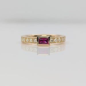 Puede incluir: Un anillo de oro con una piedra preciosa rectangular de color rosa intenso en el centro. La banda está adornada con pequeñas piedras preciosas transparentes. El anillo está sobre un fondo blanco.