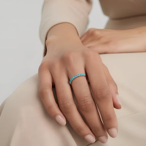 以下が含まれることがあります： 小さな明るいターコイズ色の石が並んだゴールドリング。指にはめられ、ジュエリーのデザインと色を際立たせています。シンプルでありながらエレガントなデザインのリングは、汎用性の高いアクセサリーです。