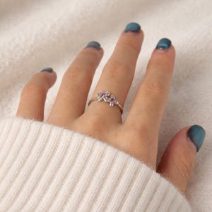 May include: A silver ring with two flower motifs, each with a pink gemstone centre. The ring is worn on a finger, set against a white sweater. The ring's delicate floral design adds a touch of elegance.