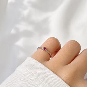May include: A silver ring with a small, round, red gemstone set in the center. The ring has a delicate, textured band with a repeating pattern. The ring is shown on a finger against a white background.