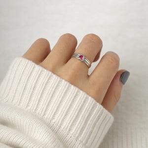 May include: A silver ring with a small, round, bright pink gemstone. The ring has a simple, elegant design with three bands. The ring is worn on a finger, and the background is a soft, white color.
