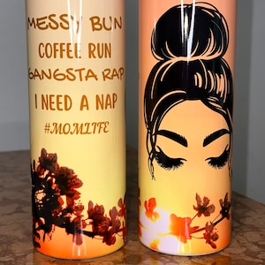 Puede incluir: Dos vasos de acero inoxidable con fondo de puesta de sol. Un vaso presenta a una mujer con un moño desordenado y el texto "Messy Bun Coffee Run Gangsta Rap I Need A Nap #MomLife". El otro vaso presenta a una mujer con un moño desordenado y pestañas largas.