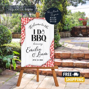 May include: A wooden easel displays a white sign with the text "Welcome to the I Do BBQ" honoring Emilia & Liam, with the date June 24, 2026. The sign is framed with red and white checkered fabric. Printed sizes are 16" x 20", 20" x 30", and 24" x 36".