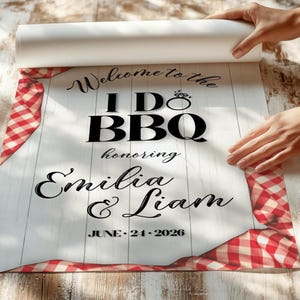 May include: A white sign with the text "Welcome to the I Do BBQ honoring Emilia & Liam JUNE 24 2026". The sign has a red and white checkered border and a faux wood grain background.