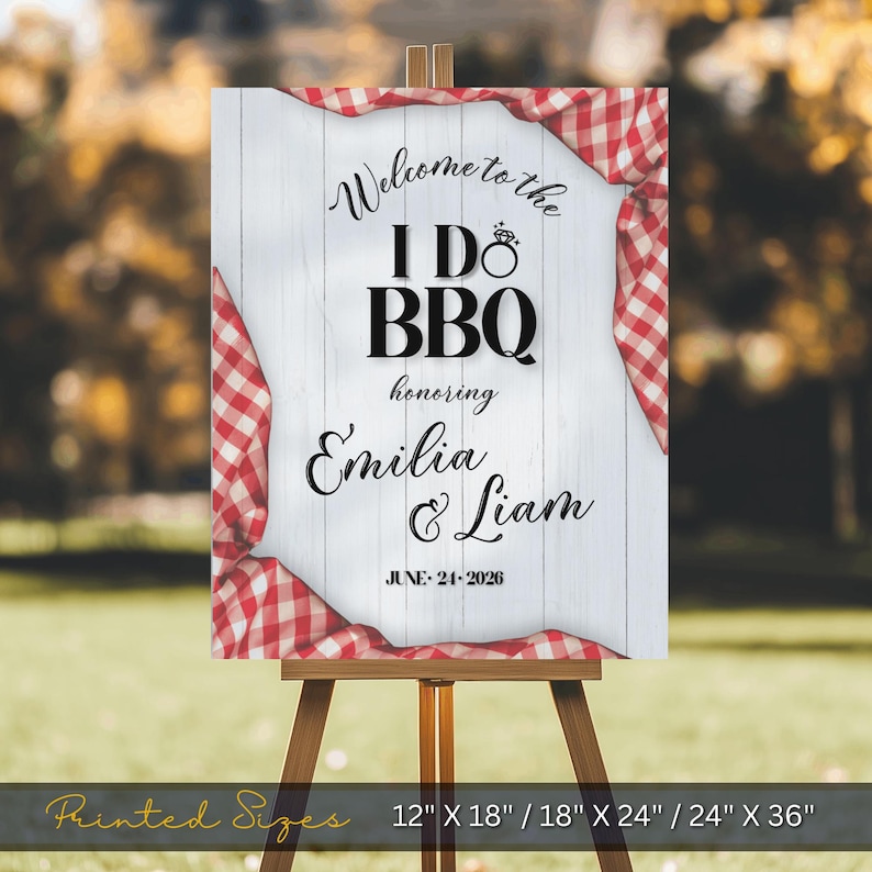 May include: A wooden easel displays a sign with the text "Welcome to the I Do BBQ" honoring Emilia & Liam, with the date June 24, 2026. The sign has a white wood background with red and white checkered fabric accents. Printed sizes are 12" X 18" / 18" X 24" / 24" X 36".
