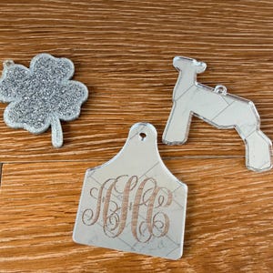 May include: Three decorative acrylic ornaments including a silver glitter four-leaf clover, a mirrored lamb silhouette, and a mirrored tag shape featuring an engraved three-letter monogram in a cursive script, each with a small hole for hanging.