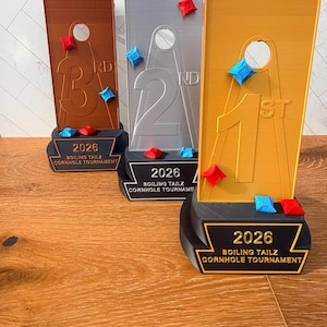 May include: Three cornhole tournament trophies in bronze, silver, and gold. Each trophy has a rectangular board with a hole and a base with the text "2026 BOILING TAILZ CORNHOLE TOURNAMENT". The boards display "1st", "2nd", and "3rd".