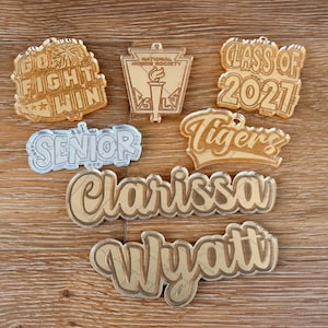 May include: A collection of acrylic graduation and school-themed charms. The charms include phrases like "Go Fight Win," "Class of 2021," and "Senior." Other charms feature names and the National Honor Society logo. The charms are in gold and silver tones.