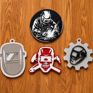 May include: A collection of welding-themed ornaments and coasters. Includes a black and white circular coaster with a welder, a gray welding mask ornament, a red and white skull ornament with crossed tools, and a white gear-shaped ornament with a welding mask.