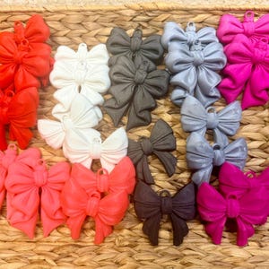 May include: A collection of colorful bow-shaped ornaments in various shades of red, pink, white, gray, and black. The bows are arranged in a woven basket, showcasing their decorative design and potential for crafting or embellishment.